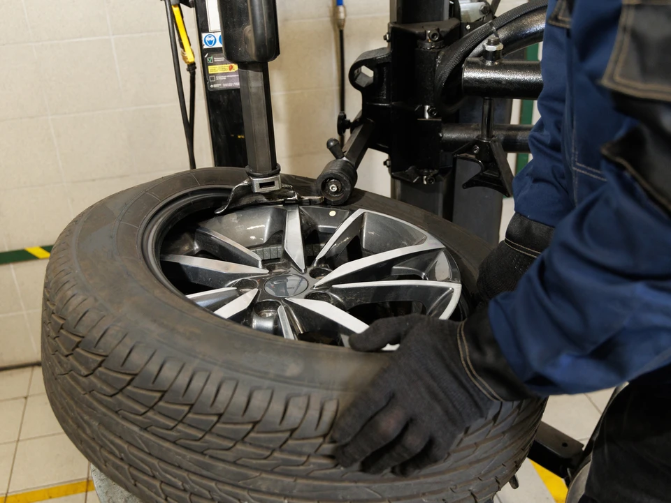 Atelier de réparation automobile avec mécanicien en combinaison grise inspectant un pneu suspendu devant un client en t-shirt blanc