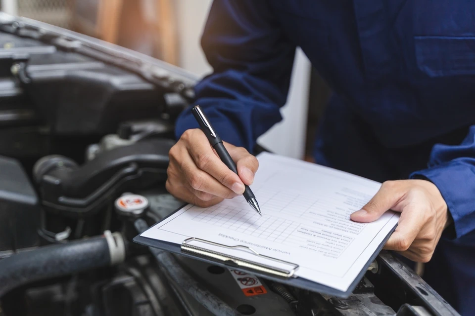 Mécanicien automobile en combinaison bleue travaillant dans un garage, consultant des documents techniques près d'un véhicule vert avec capot ouvert